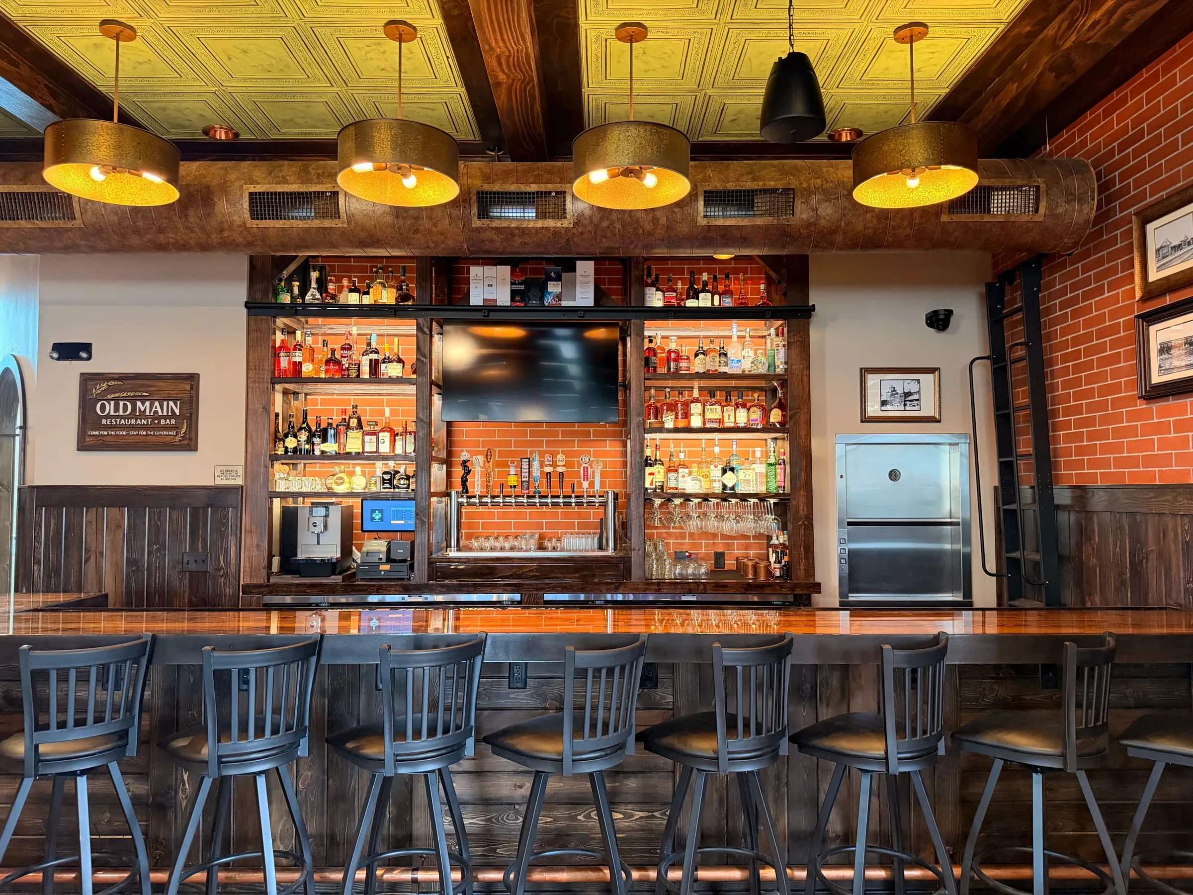 The main bar with its polished wood top and row of stools, bottles ranged across a lit back bar beneath a pressed tin ceiling and brass pendant lamps