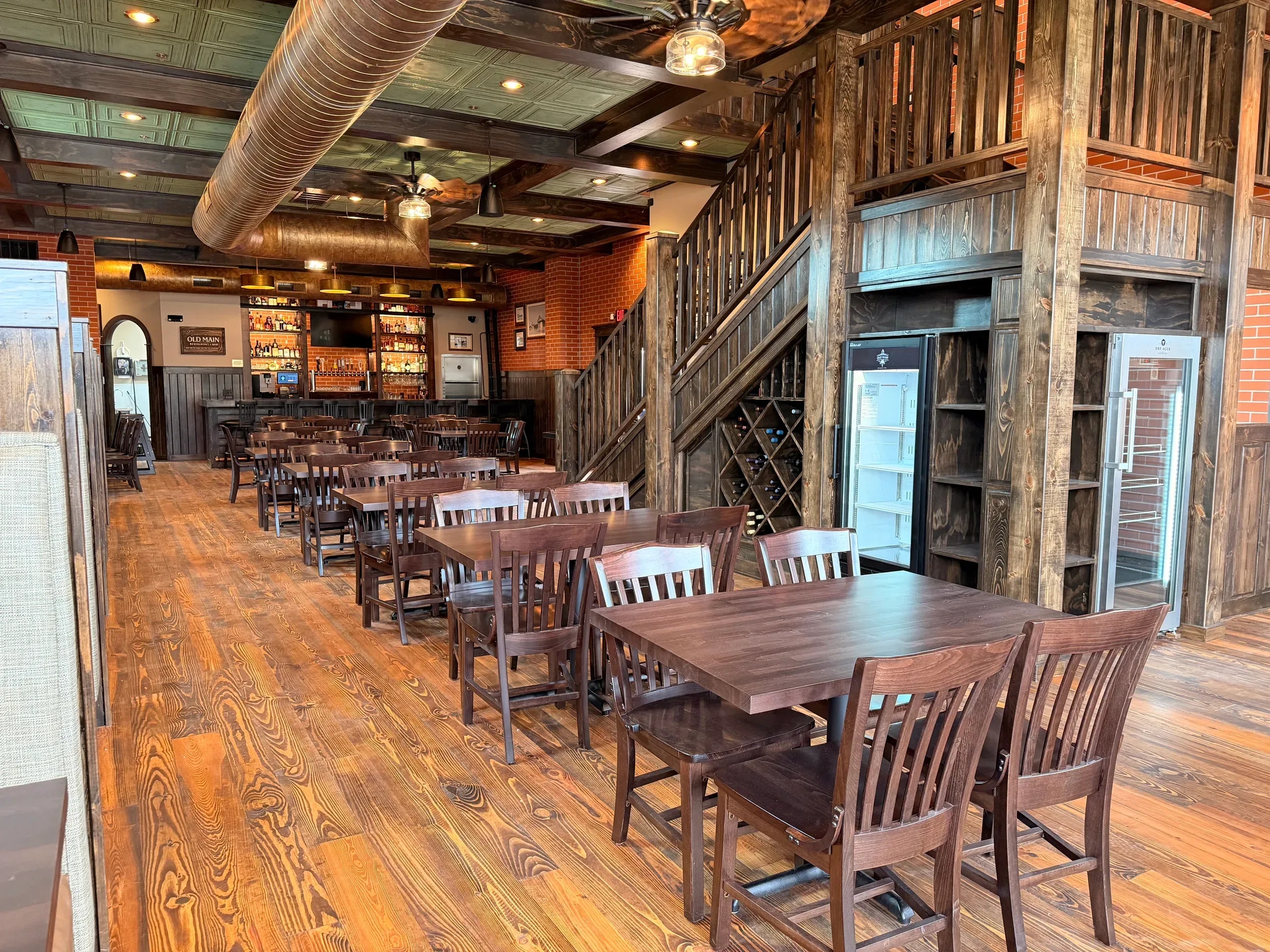 The downstairs dining room looking toward the bar, tables set along a reclaimed plank floor beneath the hand-built stair rail and diamond wine racking
