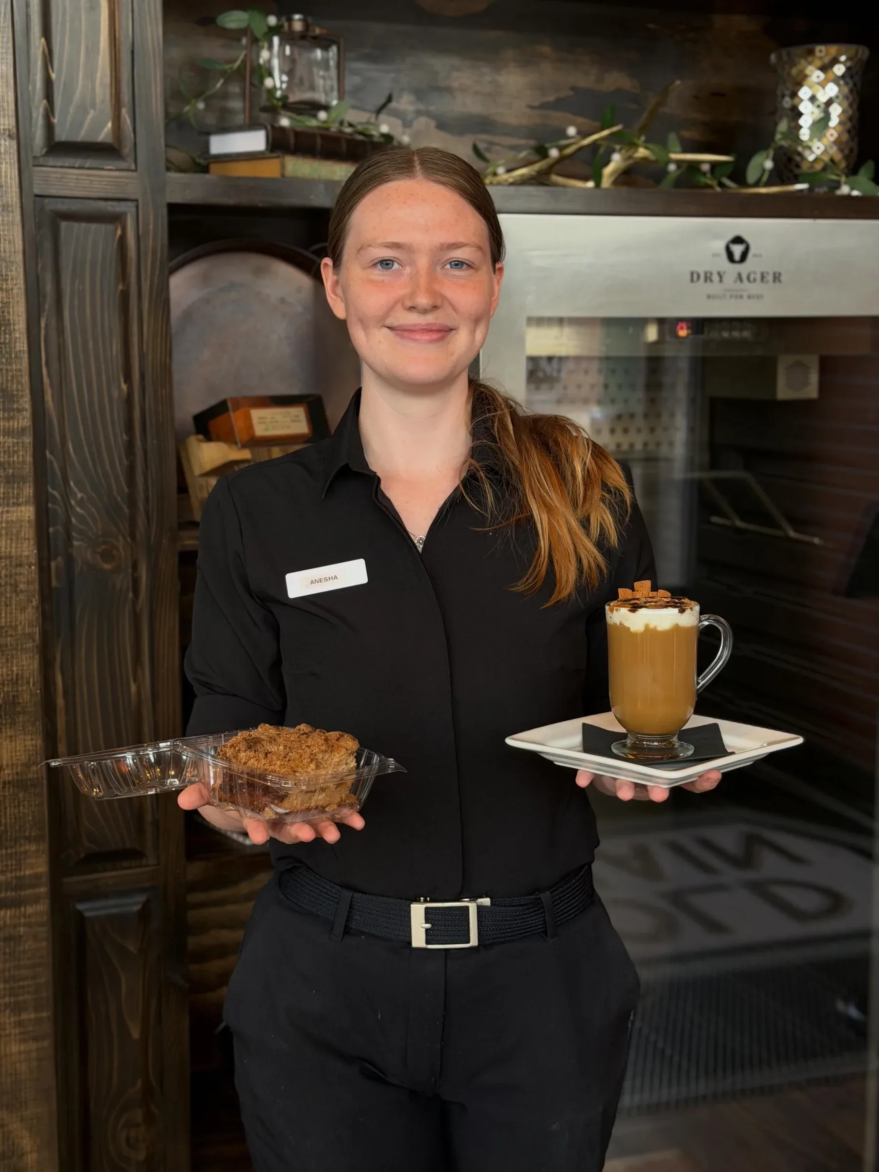A member of the Old Main team by the dry-aging cabinet just inside the front door, holding a dessert and a specialty coffee