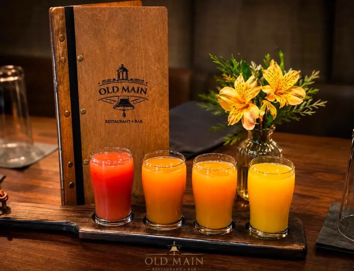 A four-glass brunch juice flight on a wooden paddle beside the engraved Old Main menu and a vase of orange lilies